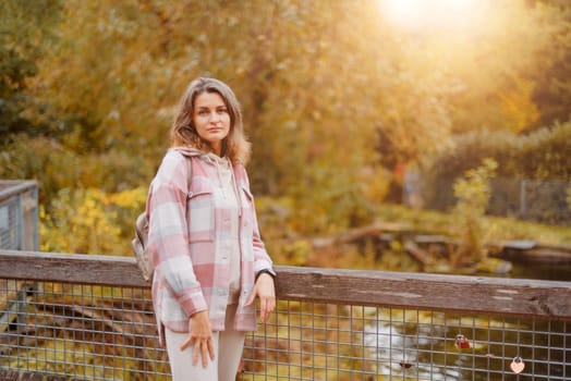 Portrait of cute young woman in casual wear in autumn, standing on bridge against background of an autumn Park and river. Pretty female walking in Park in golden fall. Copy space. smiling girl in the park standing on wooden bridge and looking at the camera in autumn season