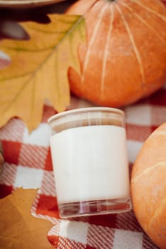White candle in transparent glass near pumpkins and bright autumn leaves
