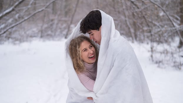 A young couple walks in the park in winter. The guy and the girl are kissing wrapped in a white blanket outdoors