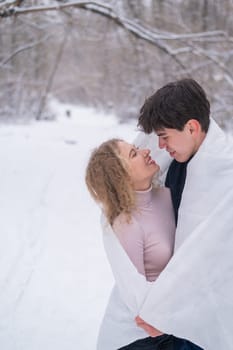 A young couple walks in the park in winter. The guy and the girl are kissing wrapped in a white blanket outdoors