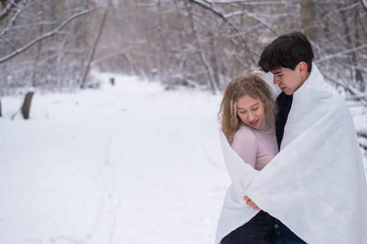 A young couple walks in the park in winter. The guy and the girl are kissing wrapped in a white blanket outdoors