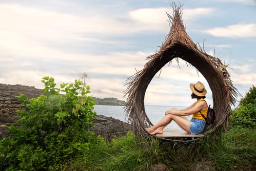 Young female traveler relaxing sitting in straw nest looking at ocean view. Woman contemplating sea landscape in Bali. Copy space. Travel, vacation concept.