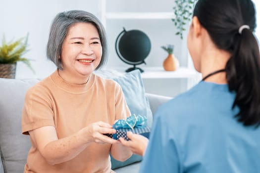 A young caregiver hand over to her senior patient a blue gift box with blue ribbons at a contented living room.