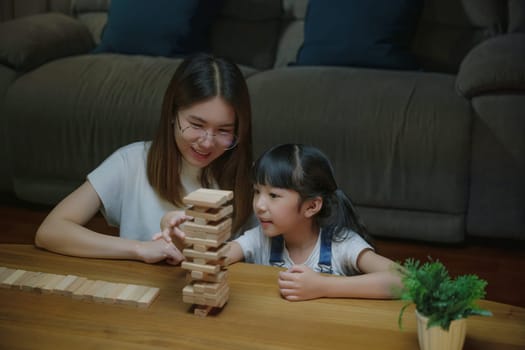 Smiling woman help teach child play build constructor of wooden blocks, Asian young mother playing game in wood block with her little daughter in home living room at night time before going to bed