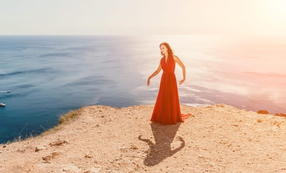 Side view a Young beautiful sensual woman in a red long dress posing on a rock high above the sea during sunrise. Girl on the nature on blue sky background. Fashion photo.