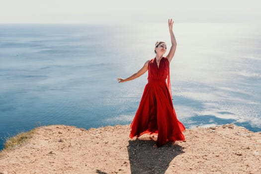 Side view a Young beautiful sensual woman in a red long dress posing on a rock high above the sea during sunrise. Girl on the nature on blue sky background. Fashion photo.