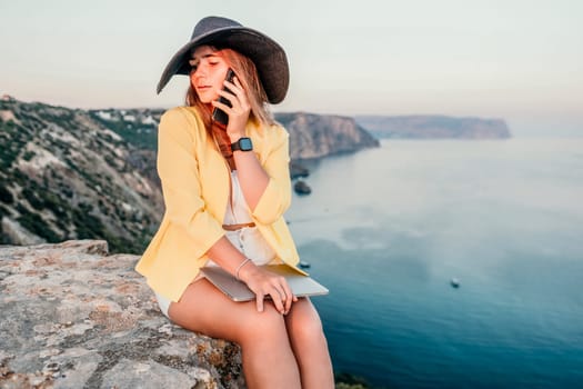 Successful business woman in yellow hat working on laptop by the sea. Pretty lady typing on computer at summer day outdoors. Freelance, travel and holidays concept.