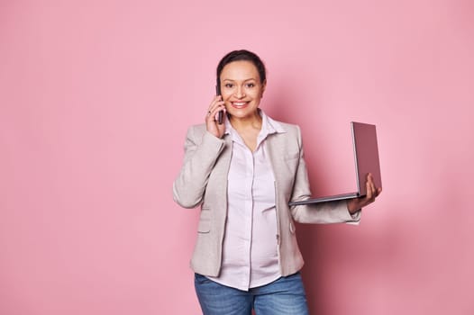 Stylish pregnant multi ethnic woman, businesswoman talking on mobile phone, negotiating with business partners, concluding deals, smiling looking at camera, posing with laptop, isolated pink backdrop