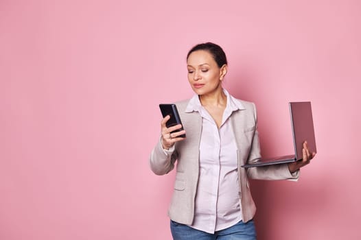 Beautiful pregnant business woman in casual clothes, holding laptop computer and smart mobile phone, looking perplexed and doubtful, isolated on pink background. People. Job. Pregnancy. Career.