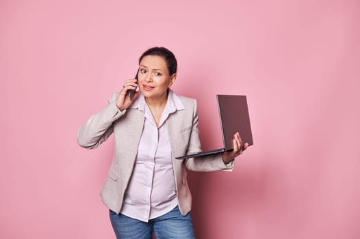 Overworked multi ethnic middle aged working pregnant business woman, holding laptop, talking on mobile phone, expressing disappointment, isolated pink background. Pregnancy. Maternity leave. Career