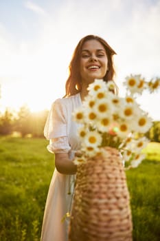 a woman in a light dress stands in a field during sunset and holds out a basket of daisies into the camera. High quality photo