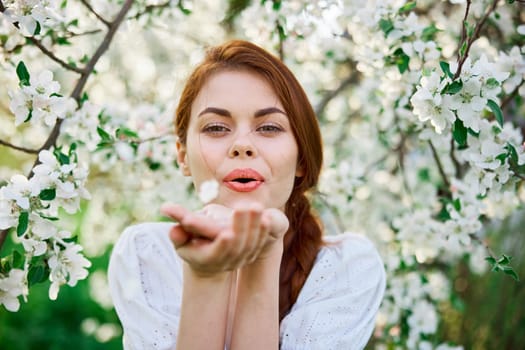 portrait of a cute girl in a light dress against the backdrop of a blooming white tree blowing flowers from her palm into the camera. High quality photo