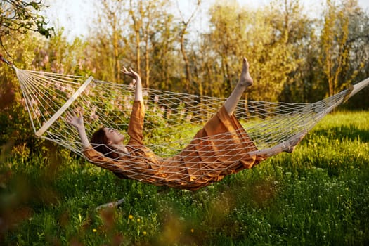 a funny woman is resting in nature lying in a mesh hammock in a long orange dress, lifting up her arms and legs, enjoying the rays of the setting sun. High quality photo