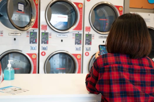 Rear view of woman sitting with mobile phone waiting for laundry washed in automatic washing machine at laundromat shop. Row of industrial washing machine in the coin laundry public laundromat.