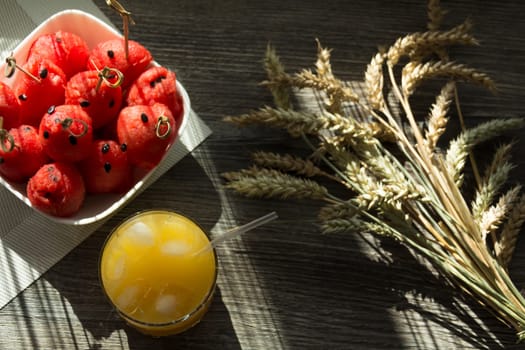 Freshly squeezed orange juice, sweet watermelon dessert and spikelets of ripe wheat on a wooden table in the sunlight.