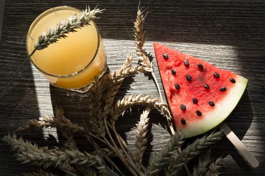 Freshly squeezed orange juice, sweet watermelon dessert and spikelets of ripe wheat on a wooden table in the sunlight
