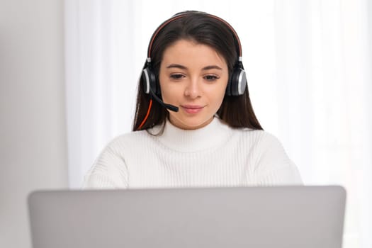 Brunette coach in headset conducts online lesson via modern laptop. Young woman sits in room speaking with students closeup