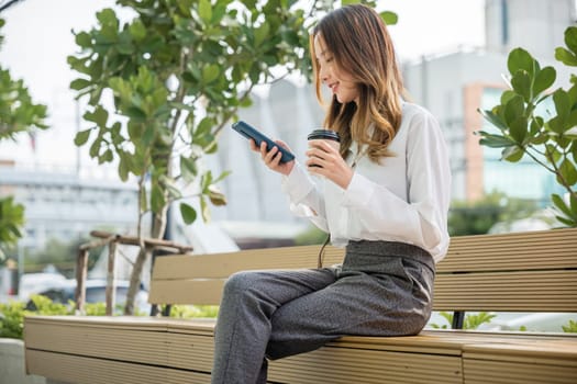Smiling woman sitting on bench outdoor on the park using smartphone chatting social media, Happy Asian businesswoman using mobile phone, technology communication, coffee paper cup takeaway