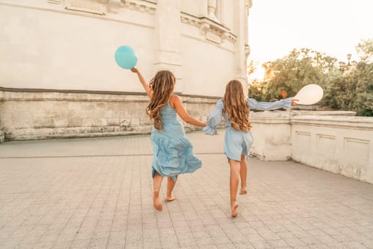 Daughter mother run holding hands. In blue dresses with flowing long hair, they hold balloons in their hands against the backdrop of a sunset and a white building