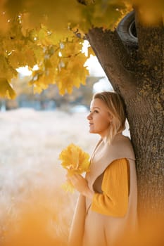 The blonde stands near the autumn tree. Thoughtful woman looks ahead, dressed in a yellow dress. Autumn content.