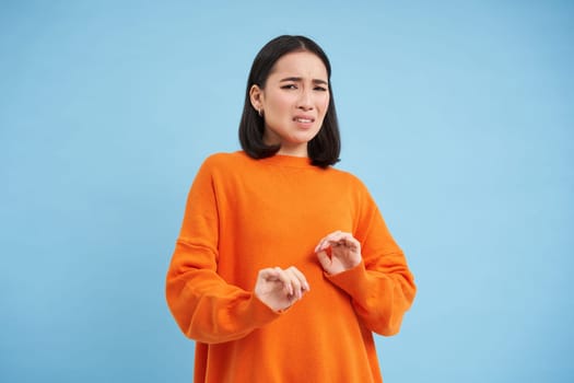 Disgusting. Asian korean girl cringes, grimaces from aversion and dislike, blocks her hands, steps back from disgust, stands over blue background.