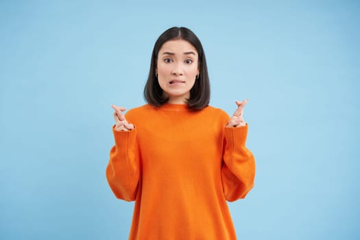 Portrait of asian woman cross fingers, hopes and prays for something, makes a wish, stands over blue background.