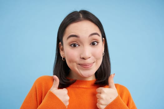 Close up portrait of asian woman shows thumbs up, smiles and looks supportive, approves, recommends smth, gives positive reply, blue background.