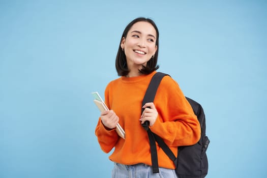 Education and students. Smiling young asian woman with backpack and notebooks, posing against blue background.