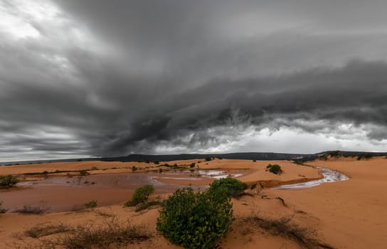 A powerful storm with black clouds rises above a desolate, distant, and gloomy sand and dune landscape.