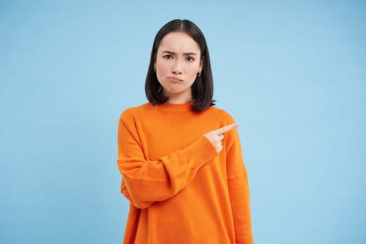 Upset brunette girl 25 years old, points right at banner, copy space on blue background, frownd with disappointed face expression.