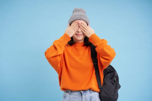 Cute asian woman with backpack, closes eyes and waits for surprise, stands over blue background. Copy space