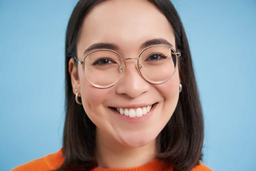 Close up portrait of beautiful young asian woman in glasses, smiling and looking happy, trying new eyewear at opticians store, stands over blue background.