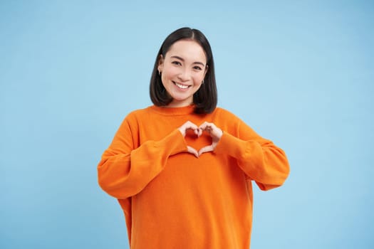 Lovely asian woman, korean girl shows heart gesture and smiles with care and tenderness, stands in orange sweatshirt against blue background.