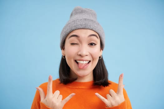 Close up portrait of funny and carefree asian girl in beanie, shows rock n roll, heavy metal gesture and tongue, stands over blue background.