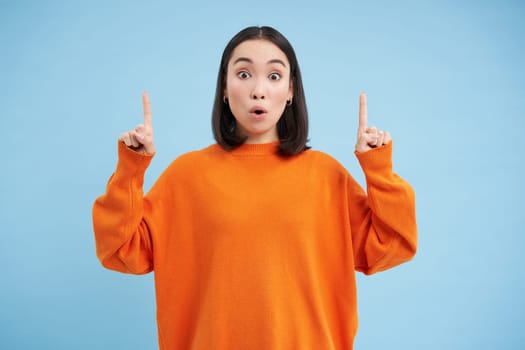 Amazed asian female model, says wow, looks impressed, points fingers up, stands over blue background. Copy space