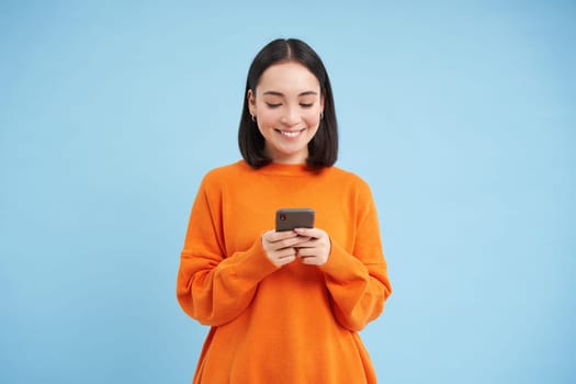 Girl with smartphone in hands smiling, using mobile application on her phone, sending message or order something online, blue background.