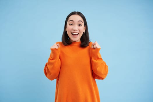 Excited young woman celebrating, jumping with hooray smiling face, winning, achieve goal, triumphing while standing over blue background.