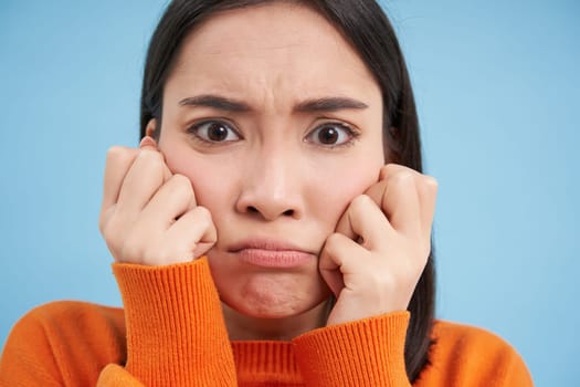 Close up of angry sulking asian woman, pouting and frowning, holding hands on cheeks, feels offended, stands over blue background.