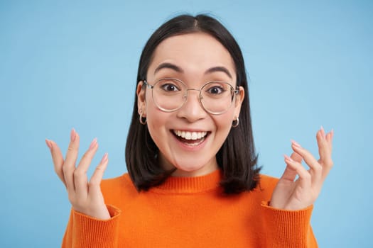 Glasses and eyewear, opticians concept. Portrait of beautiful asian woman in new spectacles, looks surprised, stands over blue background.