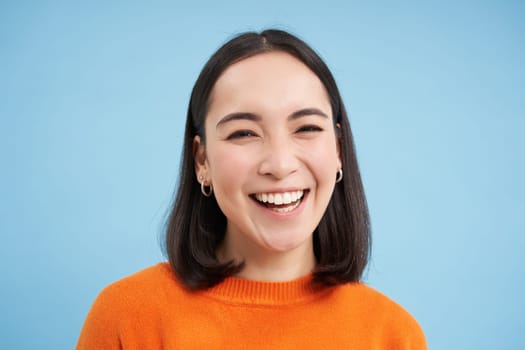 Close up of candid young asian woman, looking at camera with genuine, natural emotions, standing over blue background.