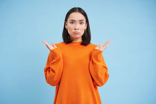 Asian girl with confused face, shrugs shoulders and pouts, looks upset and clueless, dont understand, puzzled by smth, stands over blue background.