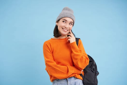 Carefree beautiful woman in streetwear, holds backpack, smiles and looks thoughtful, blue background.