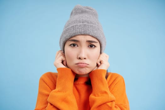 Close up portrait of korean girl feels cold, shakes and freezes, puts on beanie and sulks with sad face, stands over blue background.