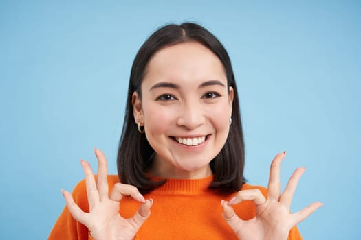Close up portrait of smiling beautiful asian woman, shows okay, ok alright sign, approves smth, approves, gives positive feedback, blue background.