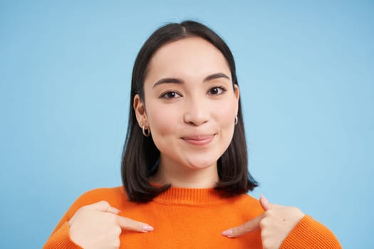 Close up of confident smiling korean girl, points at herself with pleased, satisfied face, stands over blue background. Copy space