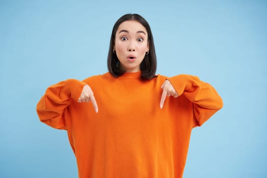 Excited asian woman pointing down, shows advertisement below with happy smile, look there gesture, stands over blue background.