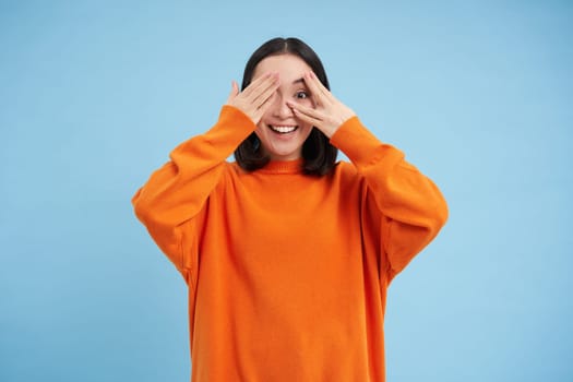 Portrait of cheerful cute asian girl waits for surprise, shuts her eyes with hands, birthday girl awaiting for gift, stands over blue background.