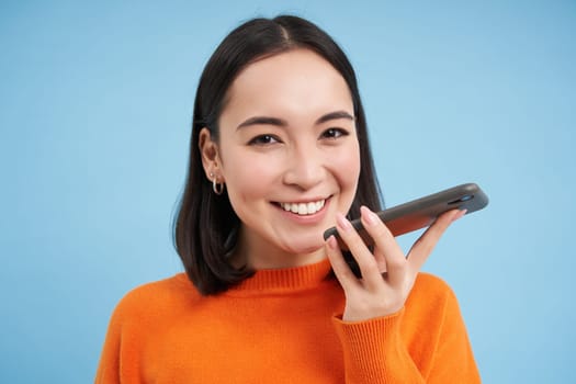 Portrait of asian woman records voice message, talks on speakerphone, holds mobile phone near mouth while speaking, translates her speech on app, blue background.