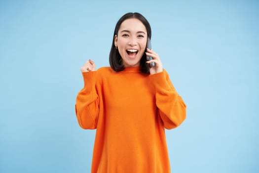 Happy asian girl receives excellent news over the phone, holds mobile and does fist pump, celebrates victory, achievement, blue studio background.