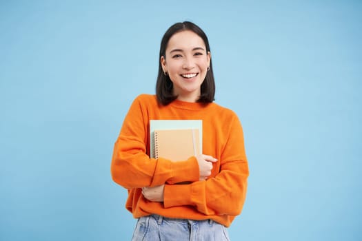 Education and students. Happy asian woman, holding notebooks and laughing, smiling at camera, enjoys going to University or College, blue background.